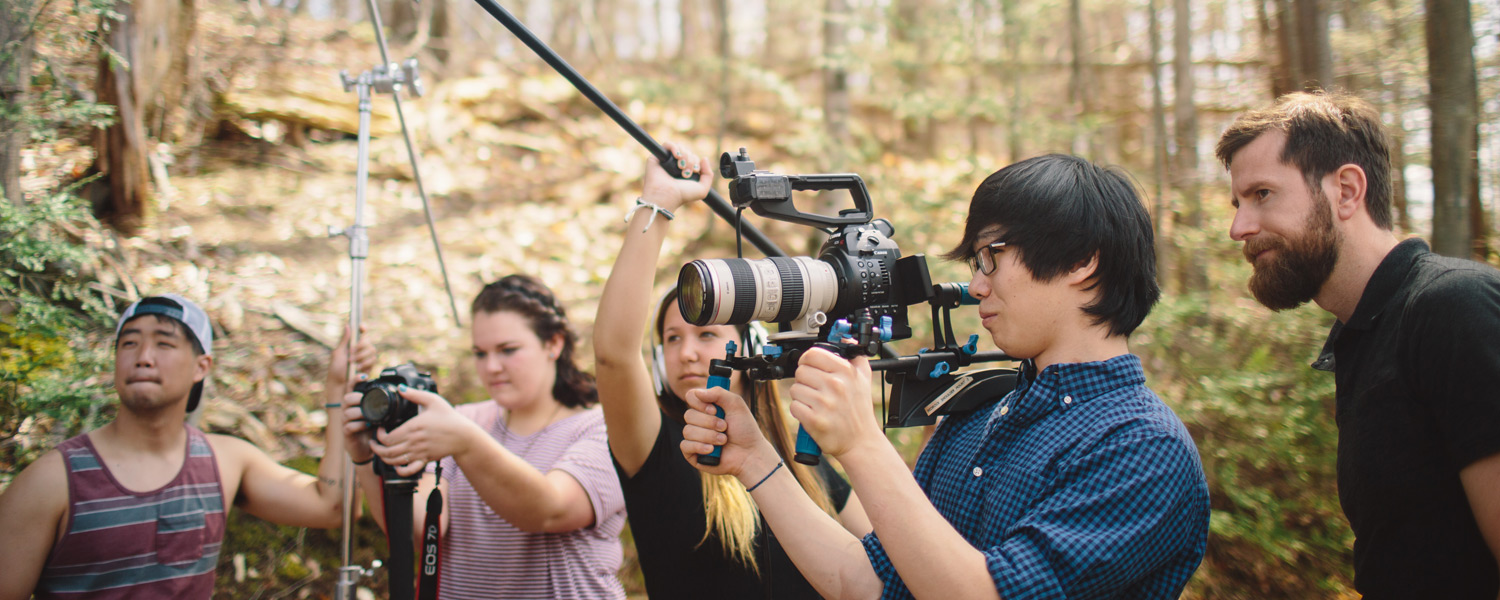 a group of Gordon college students standing in a woody outdoor area with cameras during a communication arts course at our Christian college